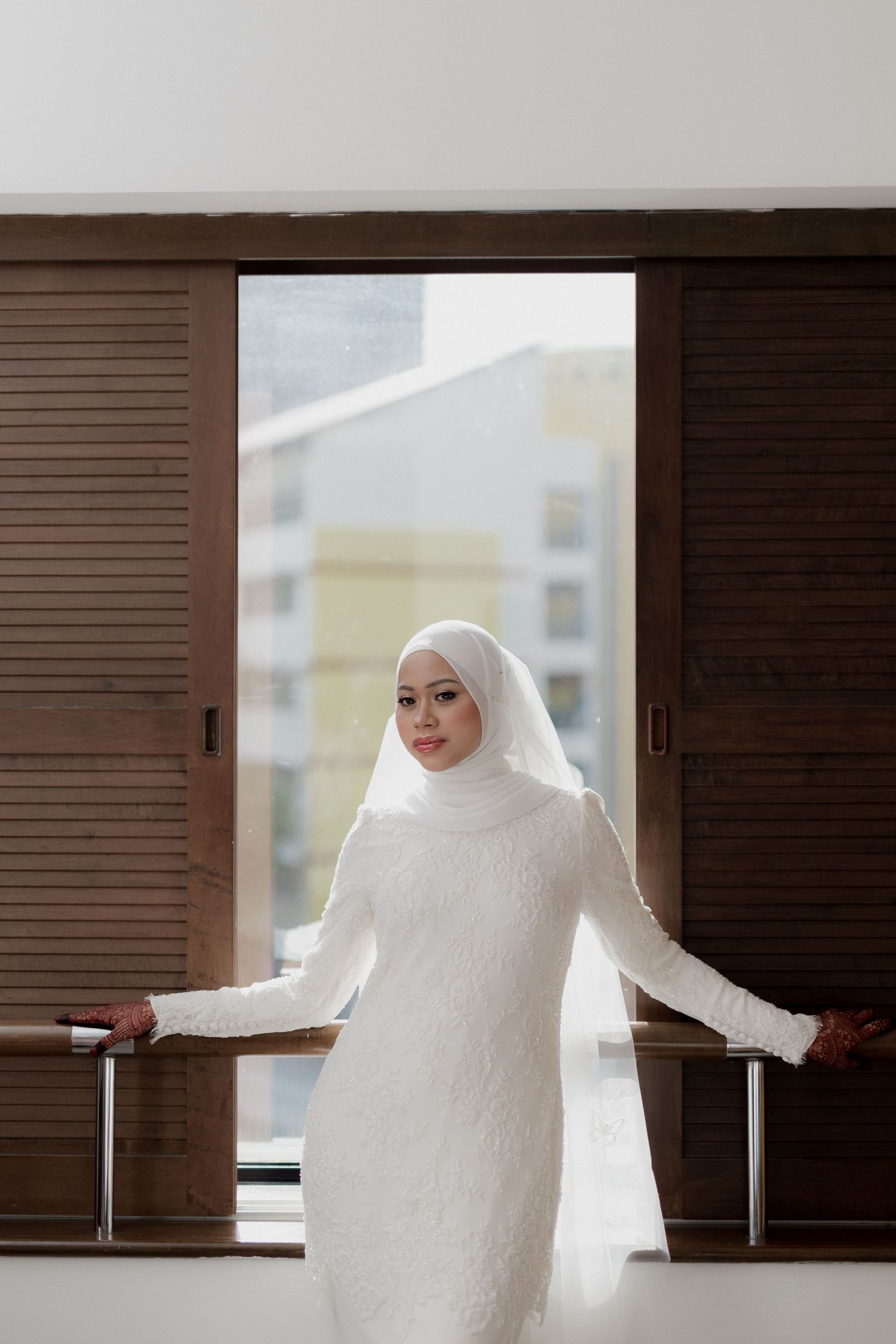 Muslim bride in white lace gown and hijab holds bouquet, walking with groom in cream suit at outdoor wedding.