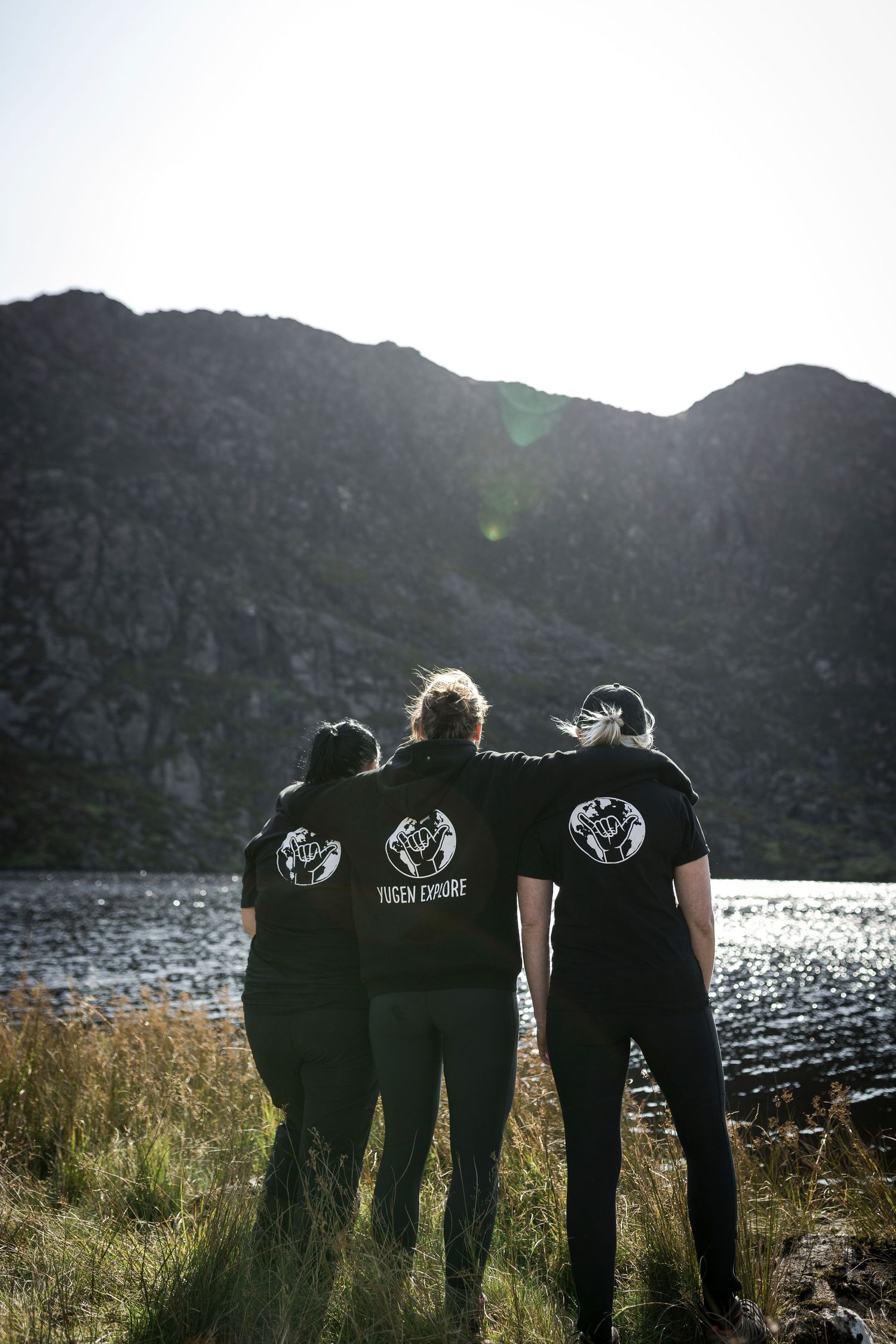Three friends in matching black Yugen Explore t-shirts stand arm in arm facing a mountain lake.