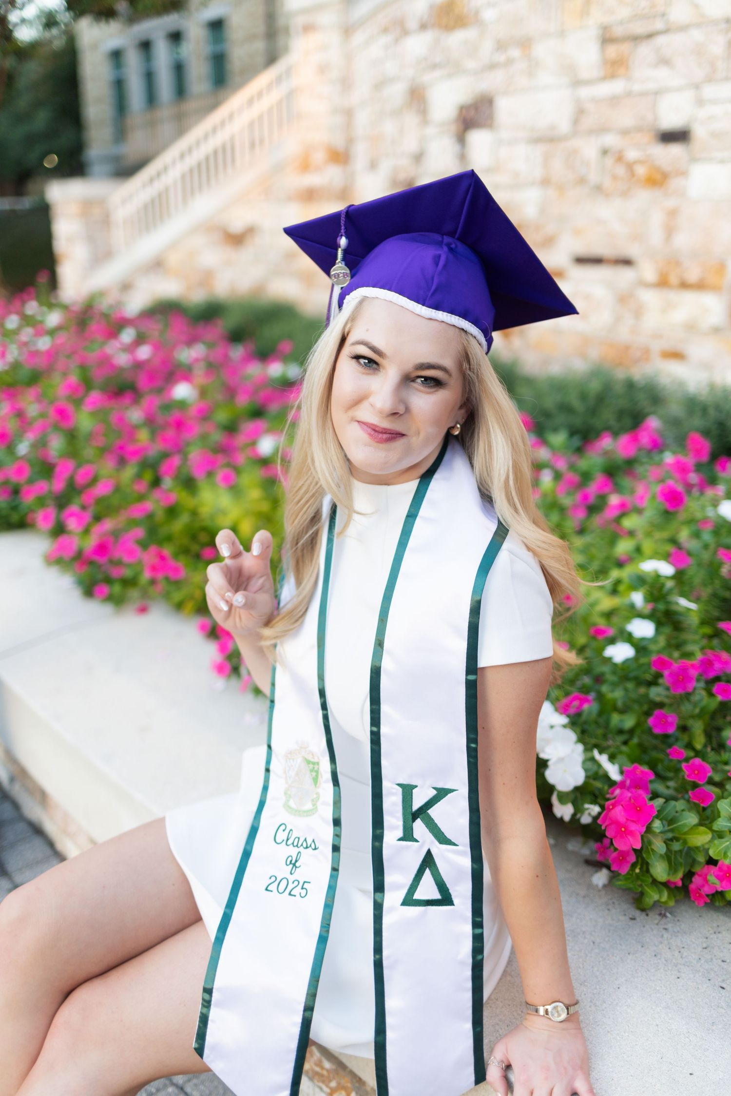 College graduate poses in purple cap and Kappa Delta stole, Class of 2025, surrounded by pink flowers.