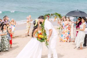 Bride and groom sharing first kiss under floral arch on Hawaiian beach.