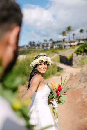 Bride in white dress and floral crown smiling over shoulder holding tropical bouquet on scenic path.