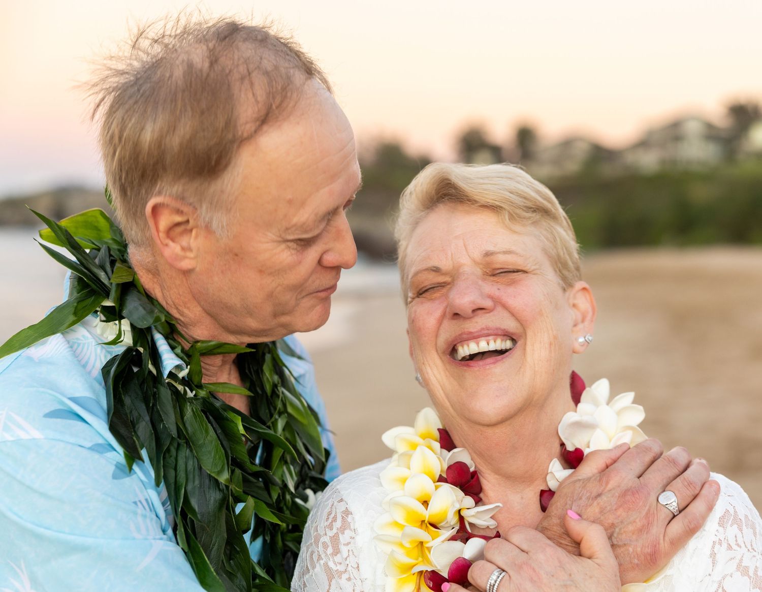 Elderly couple laughs joyfully on a Hawaiian beach wearing traditional plumeria leis at sunset.