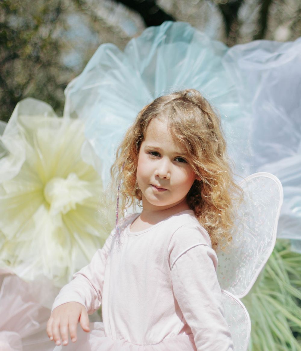 Young girl in fairy costume with white wings and pink dress, surrounded by colorful tulle decorations outdoors.