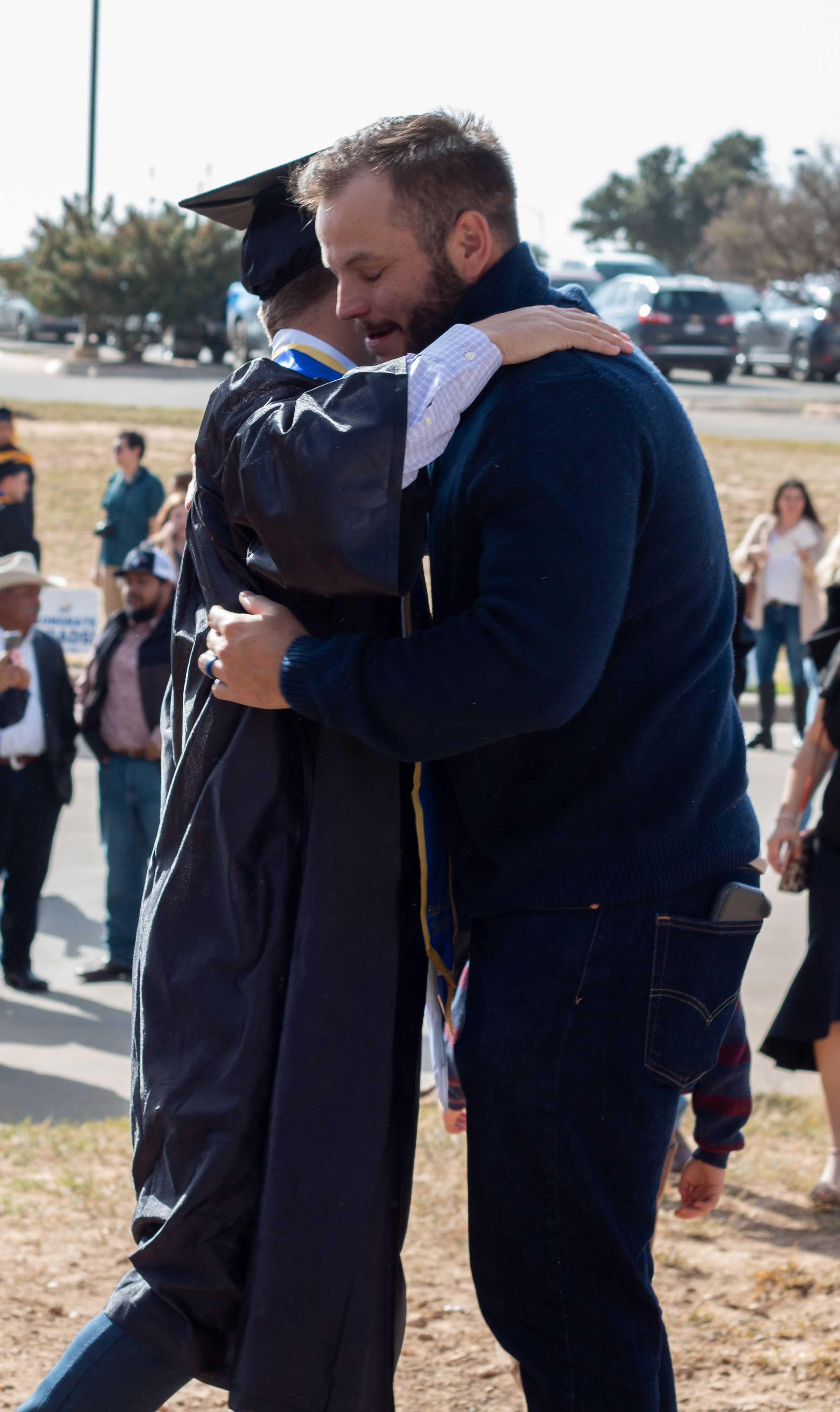 A graduate in cap and gown shares a warm hug with a bearded man in a blue jacket at an outdoor graduation ceremony.