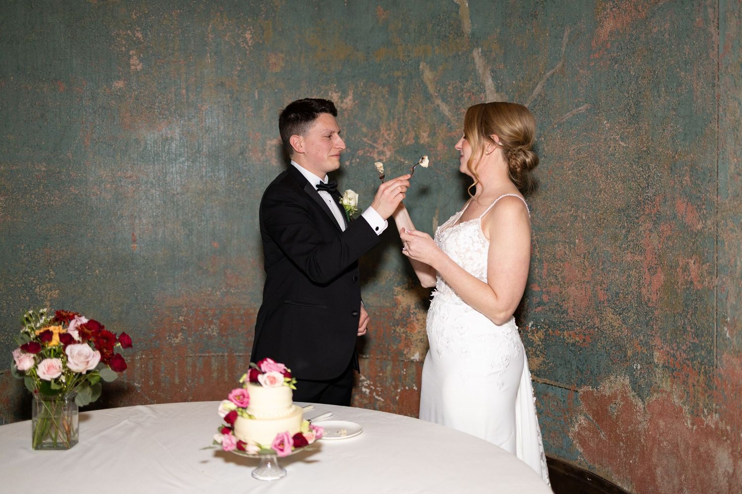 Bride and groom sharing bites of cake beside a white wedding cake at their elegant reception inside the Monastery Cincinatti wedding venue.