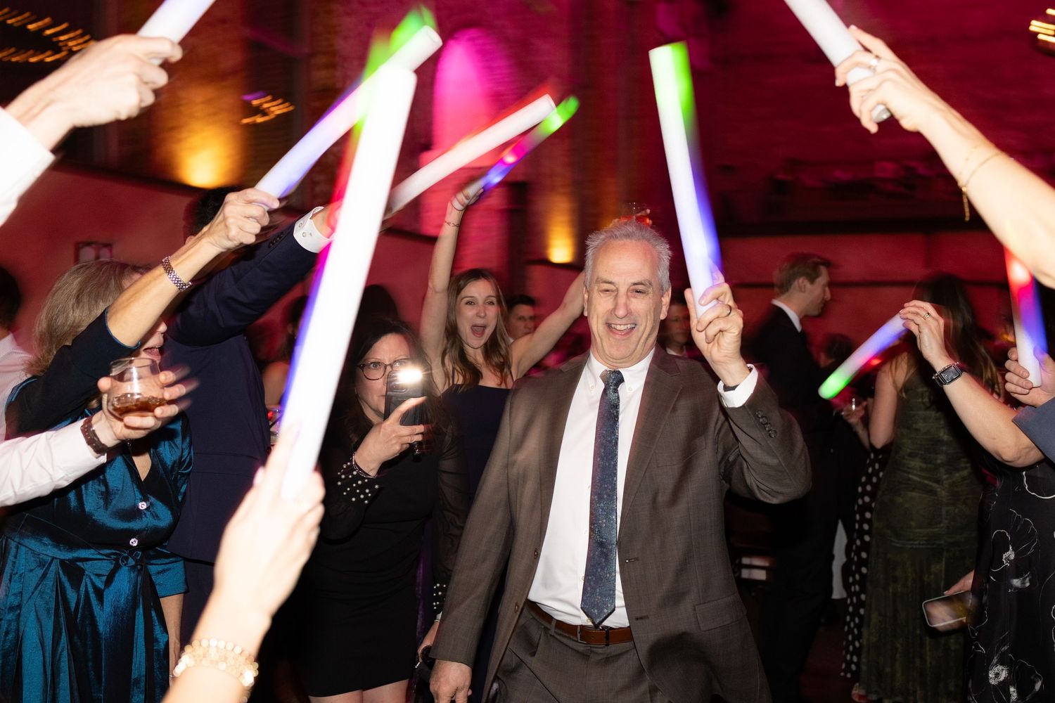 Man in suit celebrates with glowing foam sticks at a festive party event surrounded by cheering guests.