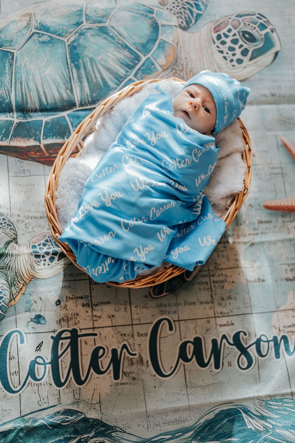 Newborn baby swaddled in blue personalized blanket and hat resting in wicker basket with Colter Carson name backdrop.