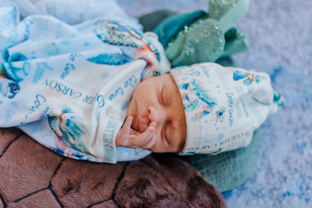 Newborn baby sleeping peacefully wrapped in a personalized blue floral swaddle blanket and matching hospital hat.
