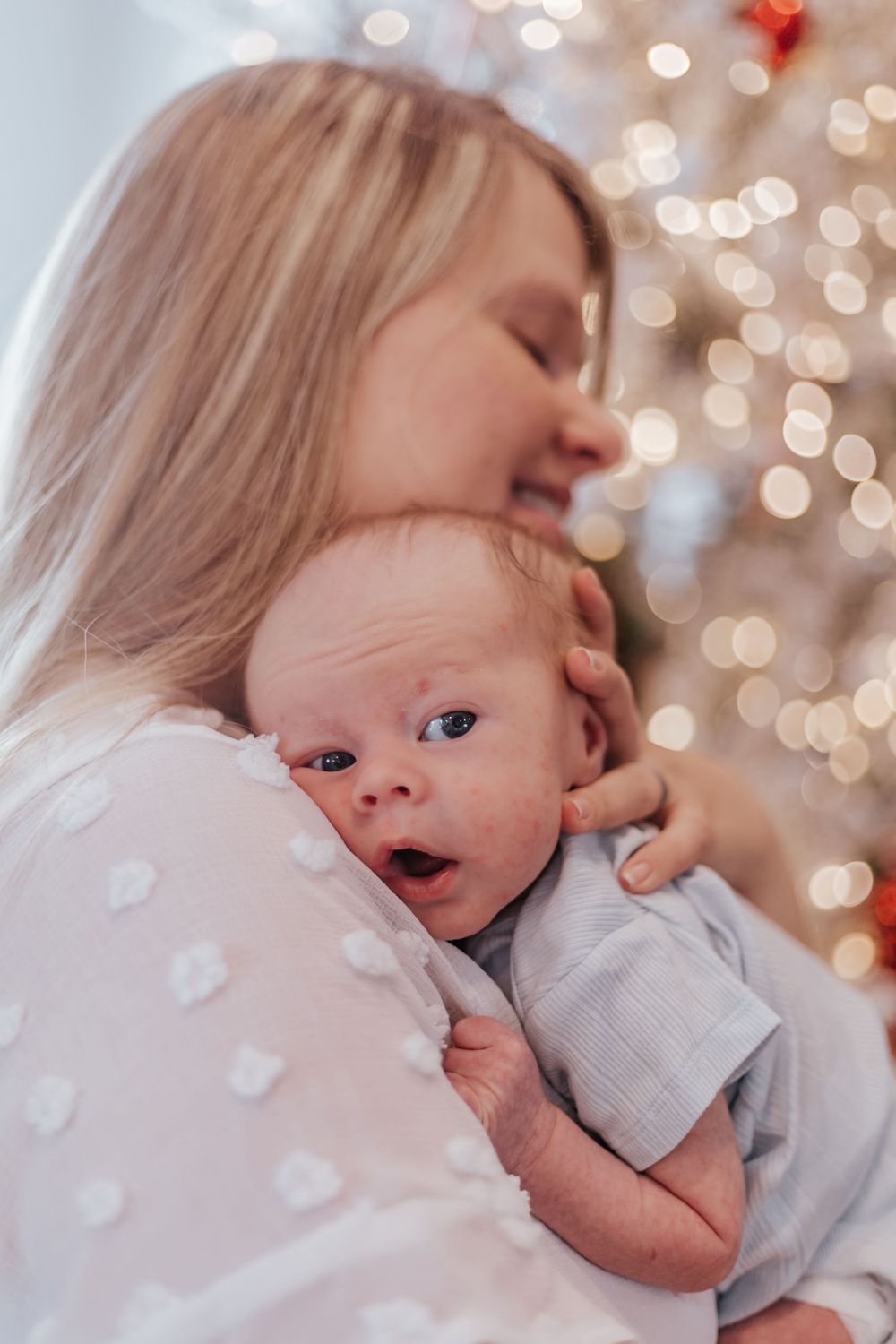 Blonde mother tenderly holding and kissing her newborn baby in front of a glowing Christmas tree.