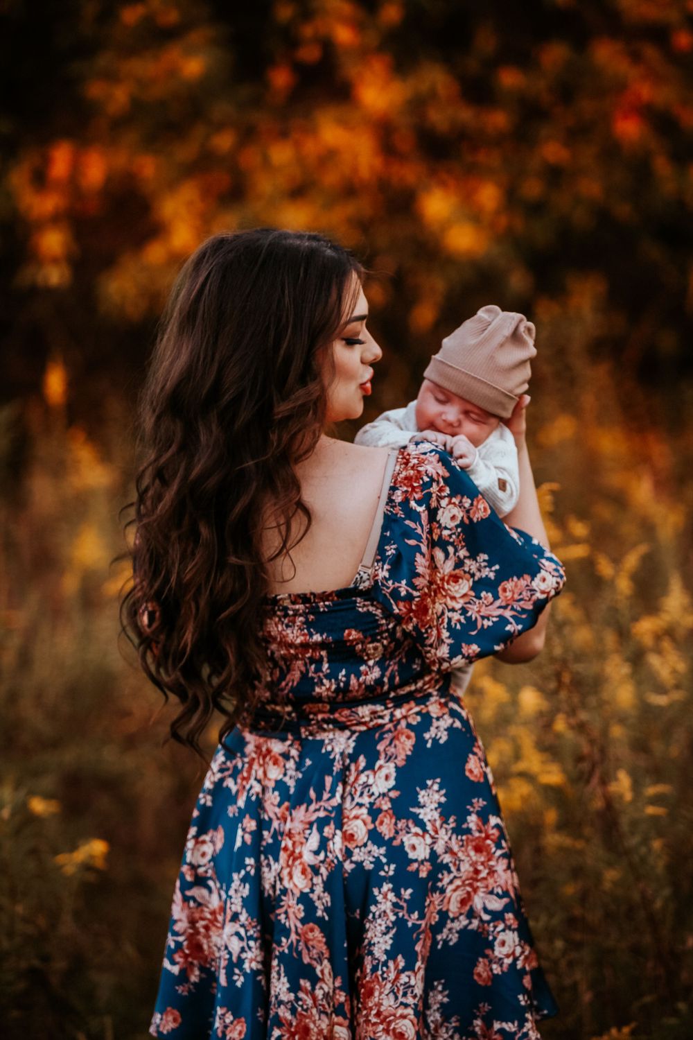 A mother in a floral blue dress kisses her newborn baby outdoors among vibrant autumn foliage.