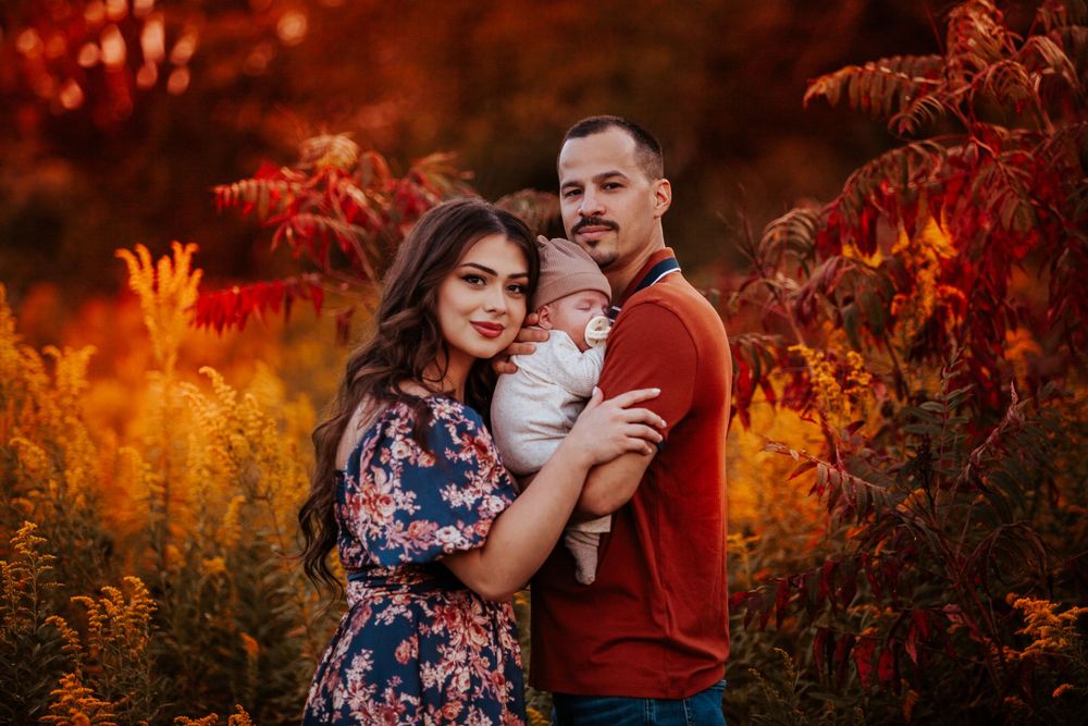 A couple holds their newborn baby outdoors surrounded by vibrant autumn foliage in warm red and orange tones.