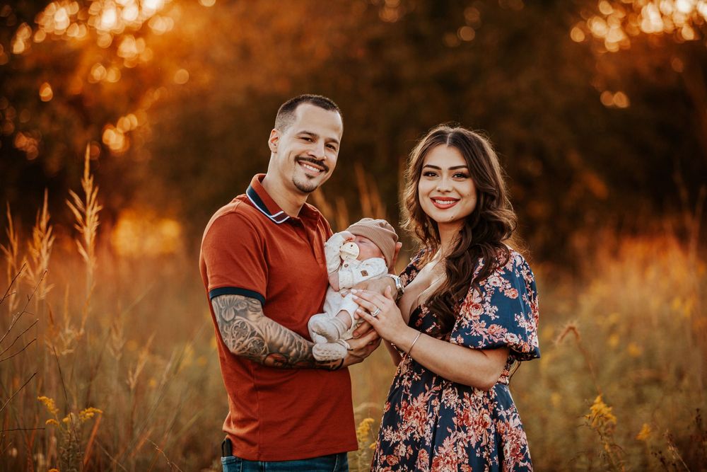 Smiling couple holding newborn baby in a sunlit autumn field with golden bokeh background.