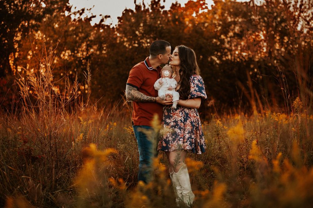 Couple kissing while holding baby in golden autumn field at sunset, woman in floral dress and white boots.