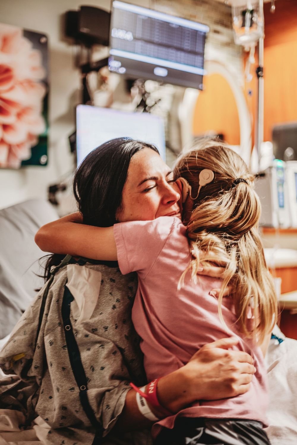A woman in a hospital gown emotionally hugs a young girl in a medical room with monitors visible in the background.