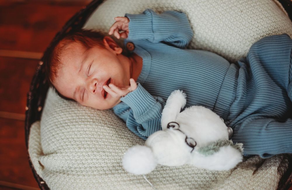 Newborn baby in blue onesie sleeping peacefully in a basket with a white stuffed bunny toy.