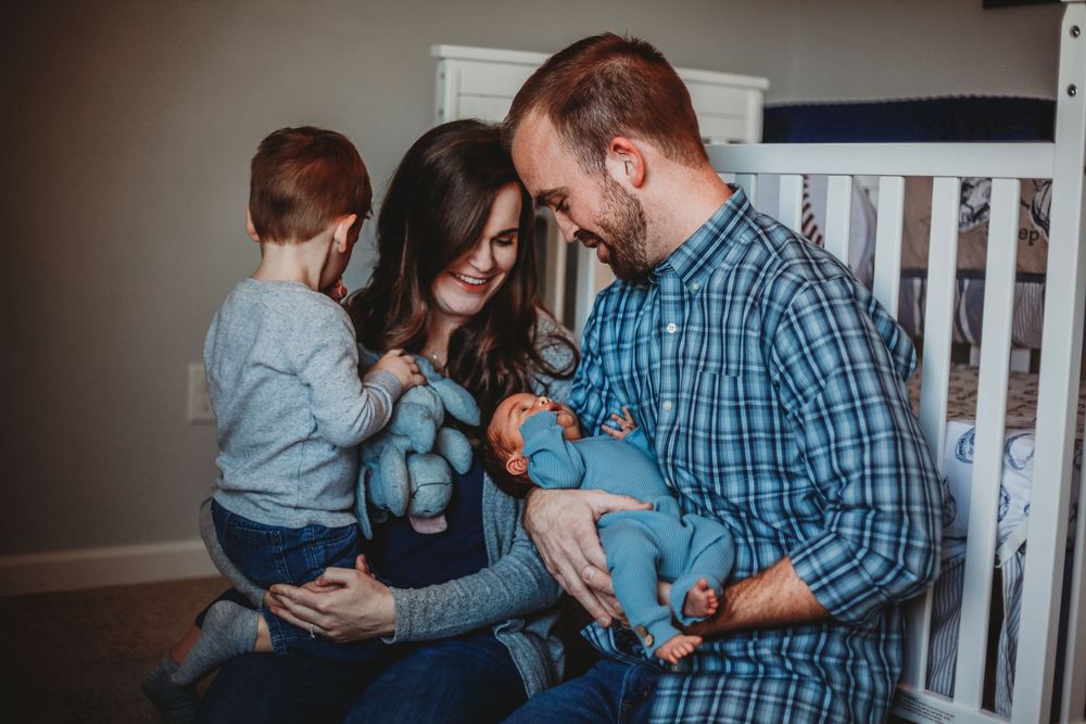 A happy family with a toddler and newborn baby poses in a nursery room for a lifestyle photo session.