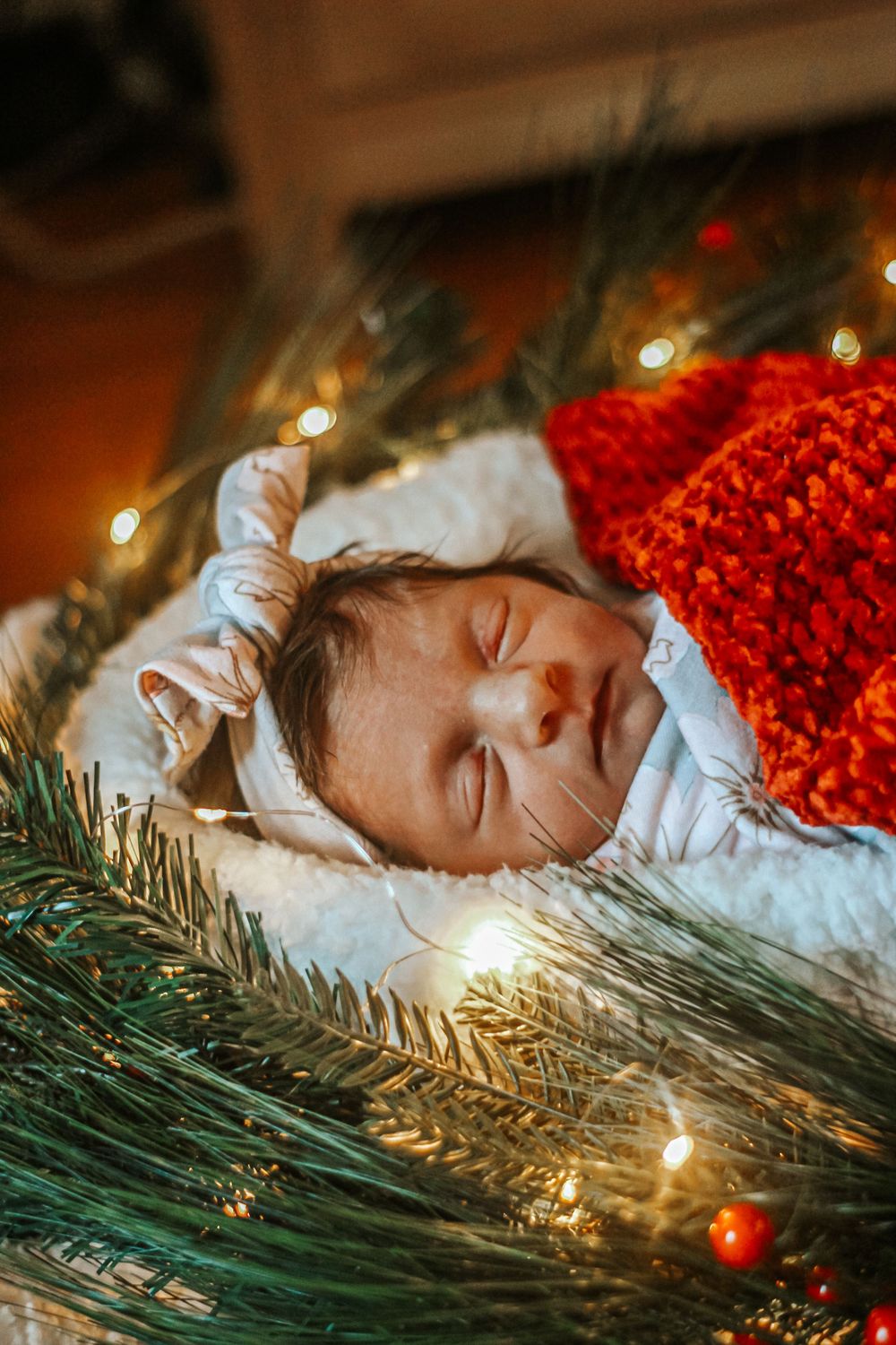 Sleeping newborn baby wrapped in red knit blanket among Christmas tree branches and twinkling fairy lights.