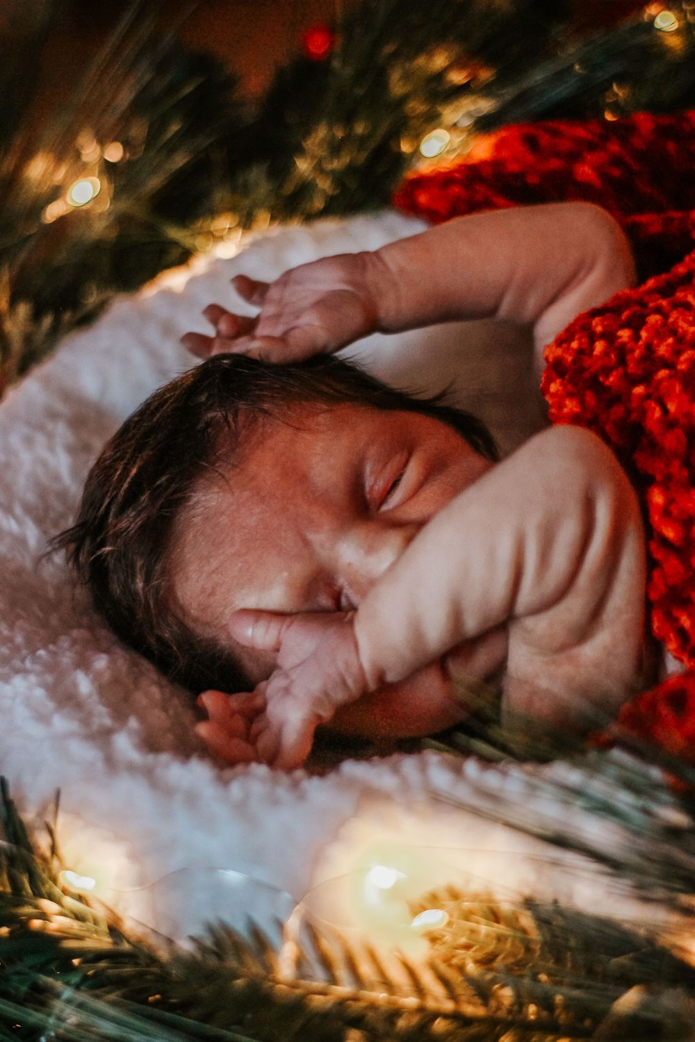 Newborn baby sleeping peacefully on white fur surrounded by Christmas tree branches and warm glowing lights.