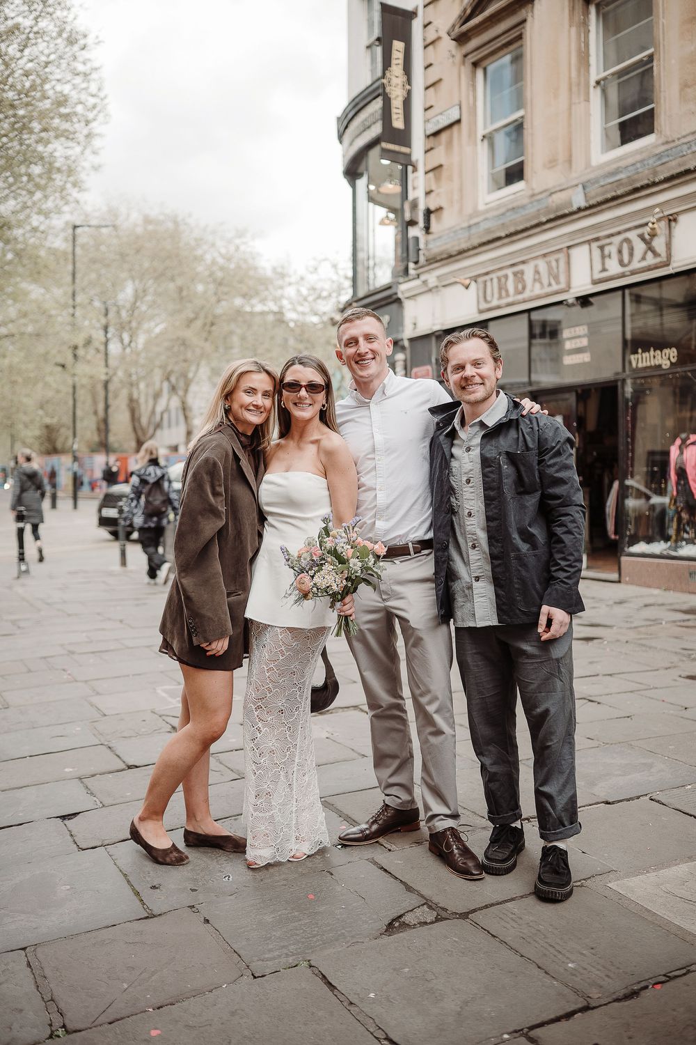Bride in white lace dress holds bouquet posing with groom and guests on a cobblestone street outside Urban Fox store.