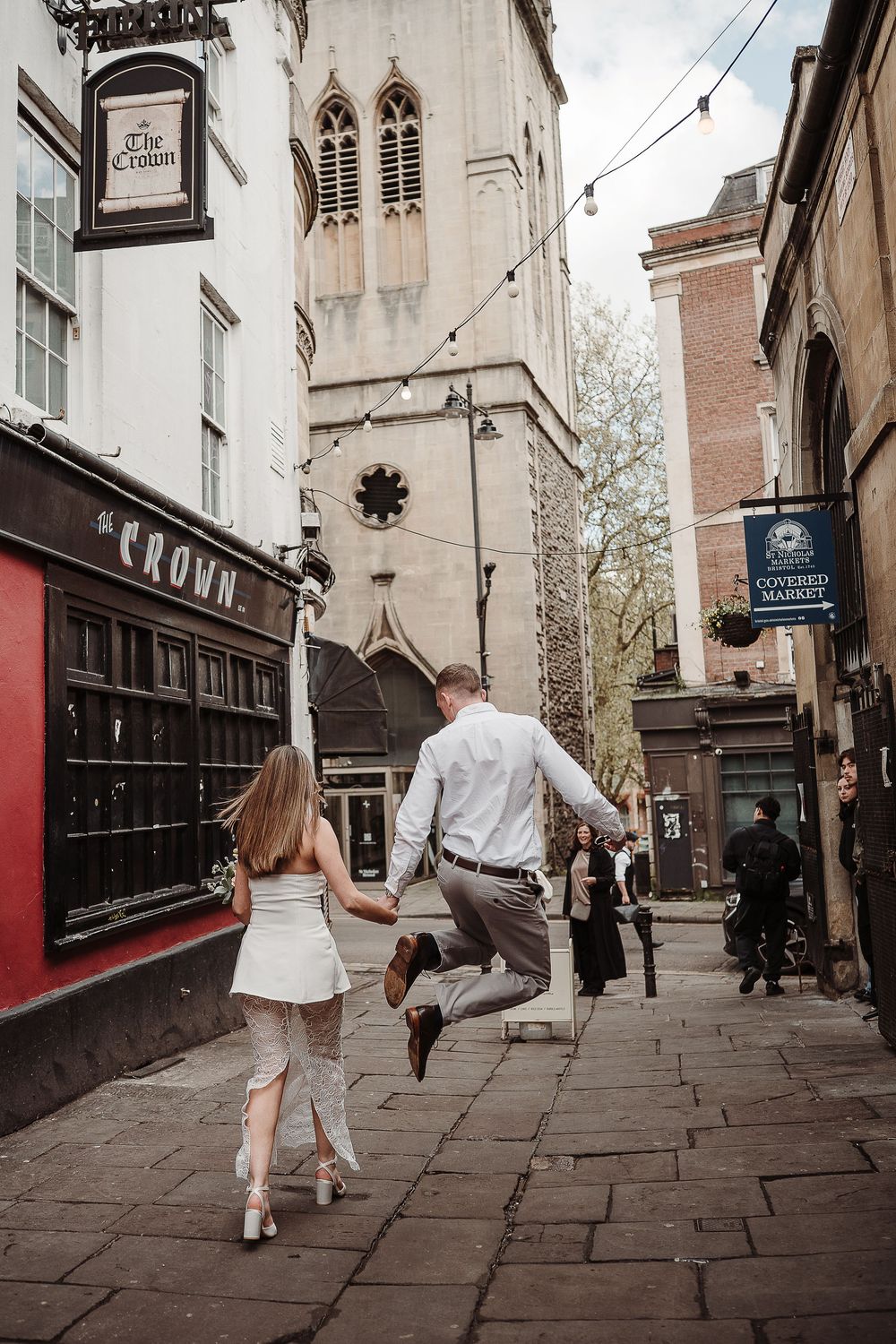 Couple holding hands and jumping joyfully on a cobblestone street in Oxford near The Crown pub and Covered Market.