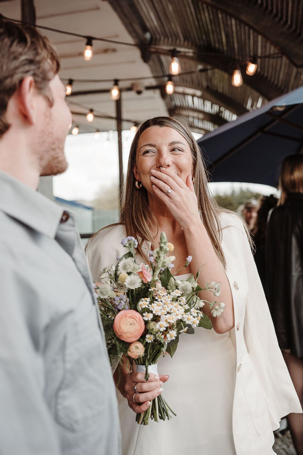 Bride in white dress laughing joyfully holding a pastel floral bouquet during an outdoor wedding celebration.