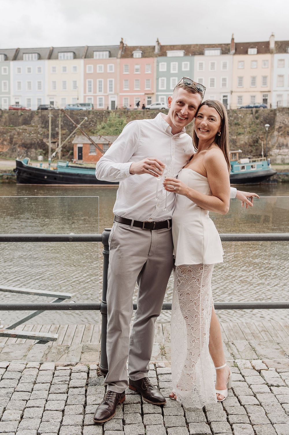 Couple posing by waterfront with colorful buildings, woman in white lace dress embracing man in white shirt and grey trousers.