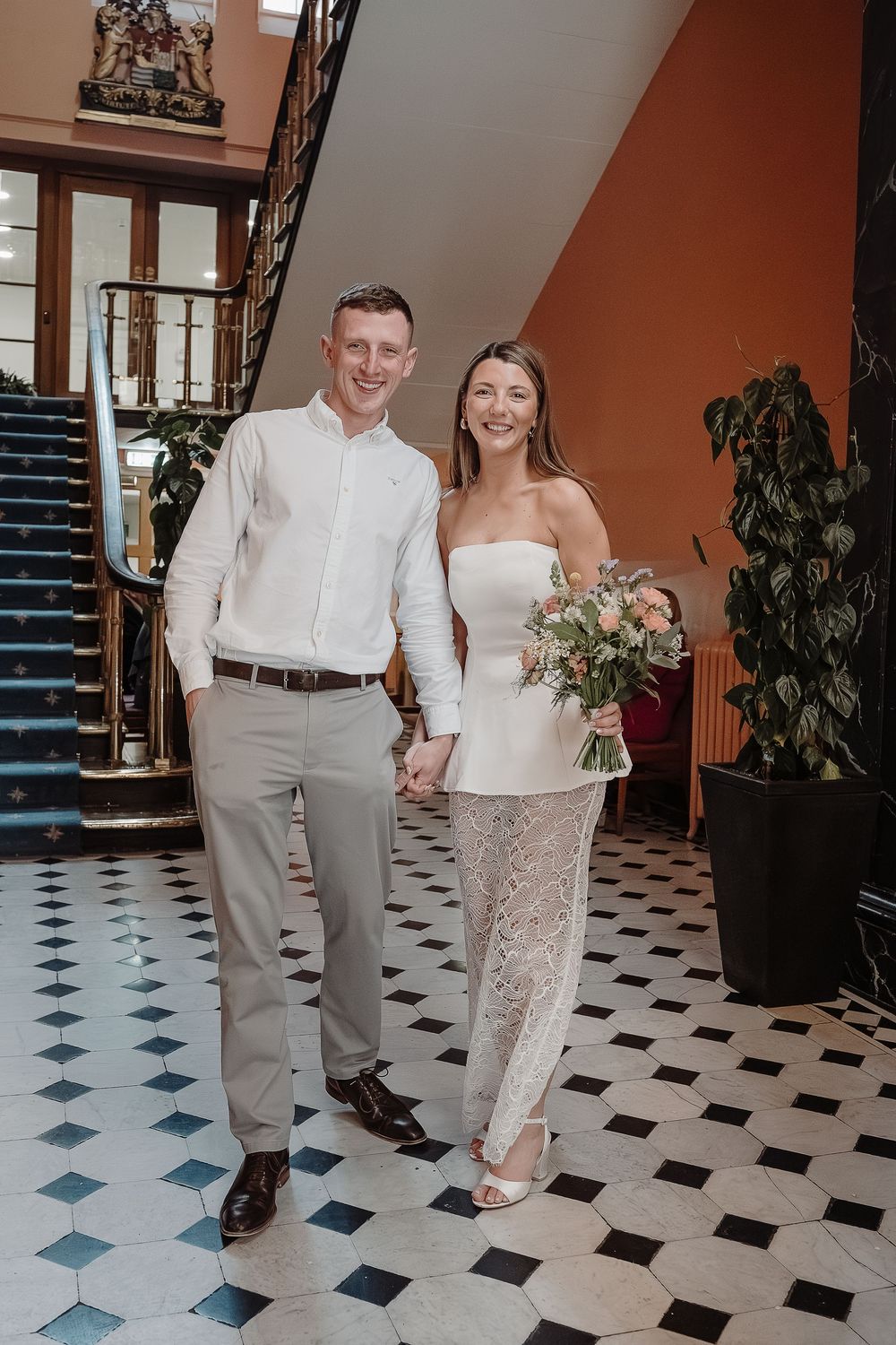 Smiling newlywed couple holding hands in elegant venue with black and white tiled floor and grand staircase.