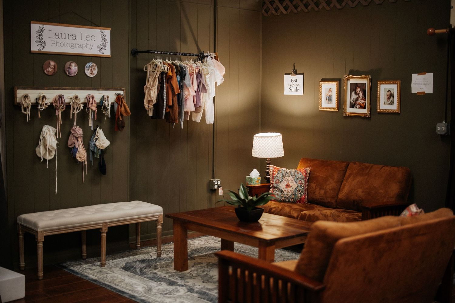 Cozy photography studio waiting room with brown leather sofa, wooden table, lamp, framed photos, and colorful newborn wraps on wall rack.