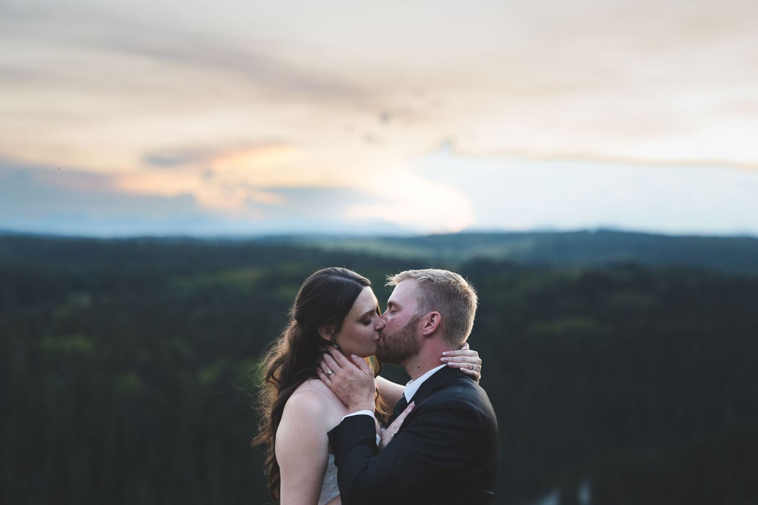 Couple sharing a romantic kiss at sunset with lush forest and dramatic sky in the background at their wedding.