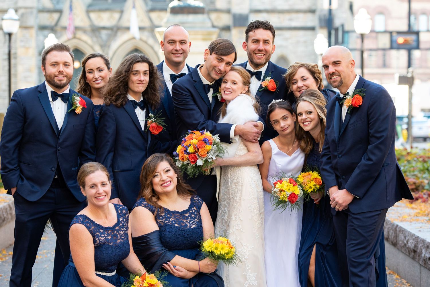 Wedding party in navy and yellow attire poses outdoors against stone building backdrop.