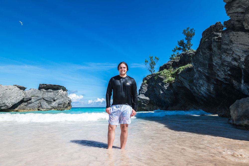 Person standing on a pink sand beach in Bermuda, wearing a black rash guard and white shorts, with rocky cliffs behind.