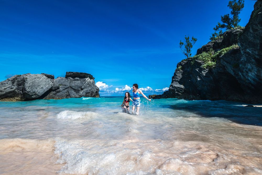Two people playing in crystal-clear turquoise waters at a rocky tropical beach under a vivid blue sky.