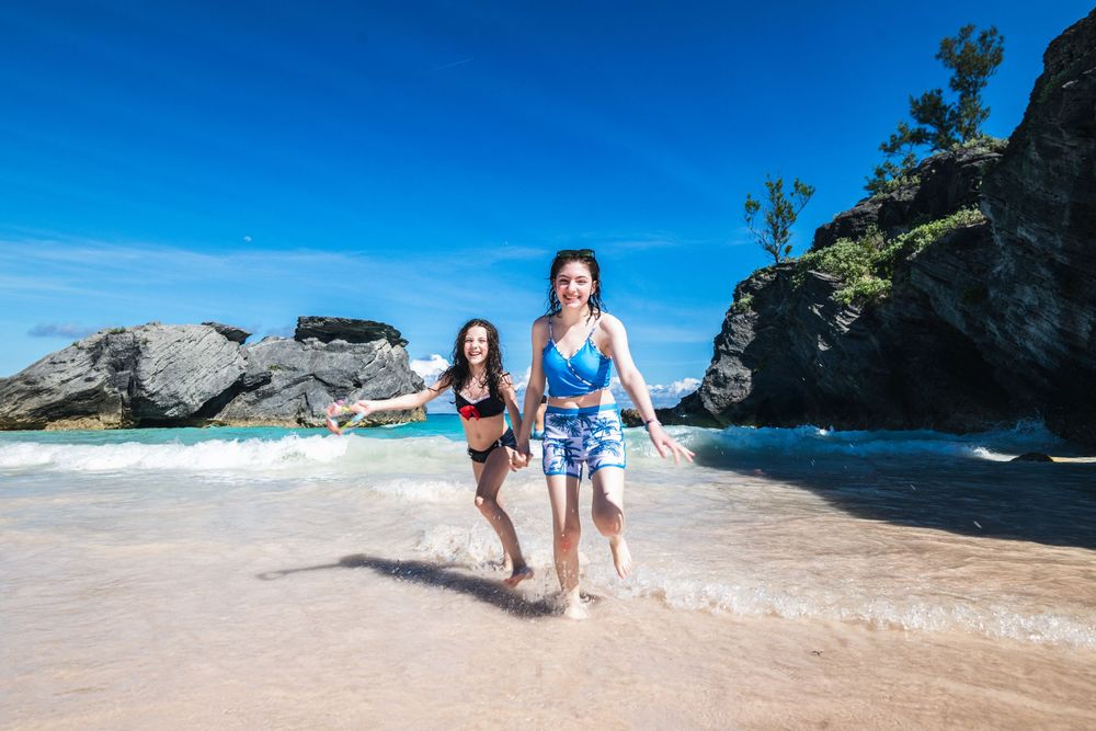 Two girls in swimsuits running and laughing on a tropical pink sand beach with rocky cliffs and blue sky.