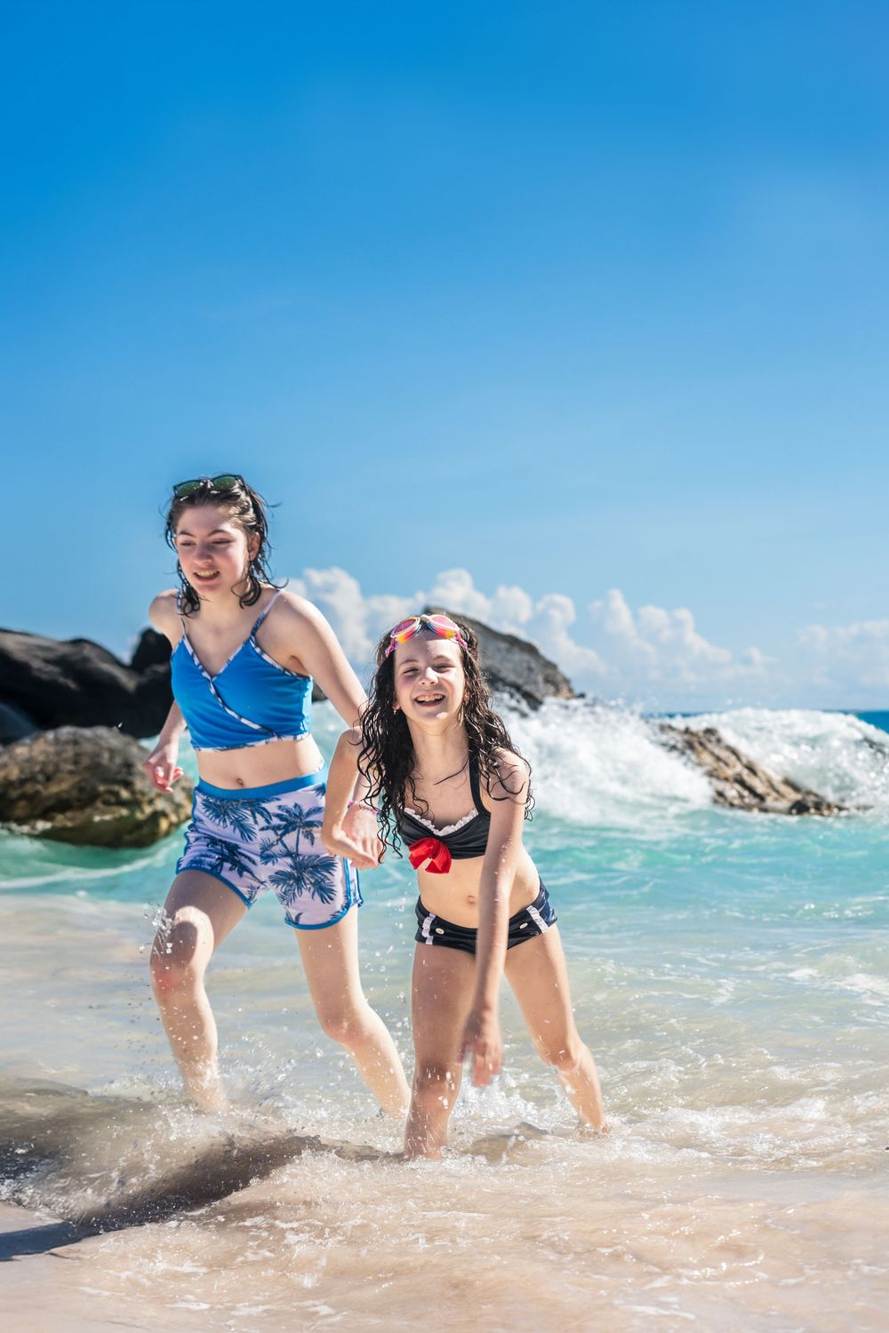 Two girls in swimsuits laughing and running through ocean waves on a sunny tropical beach.