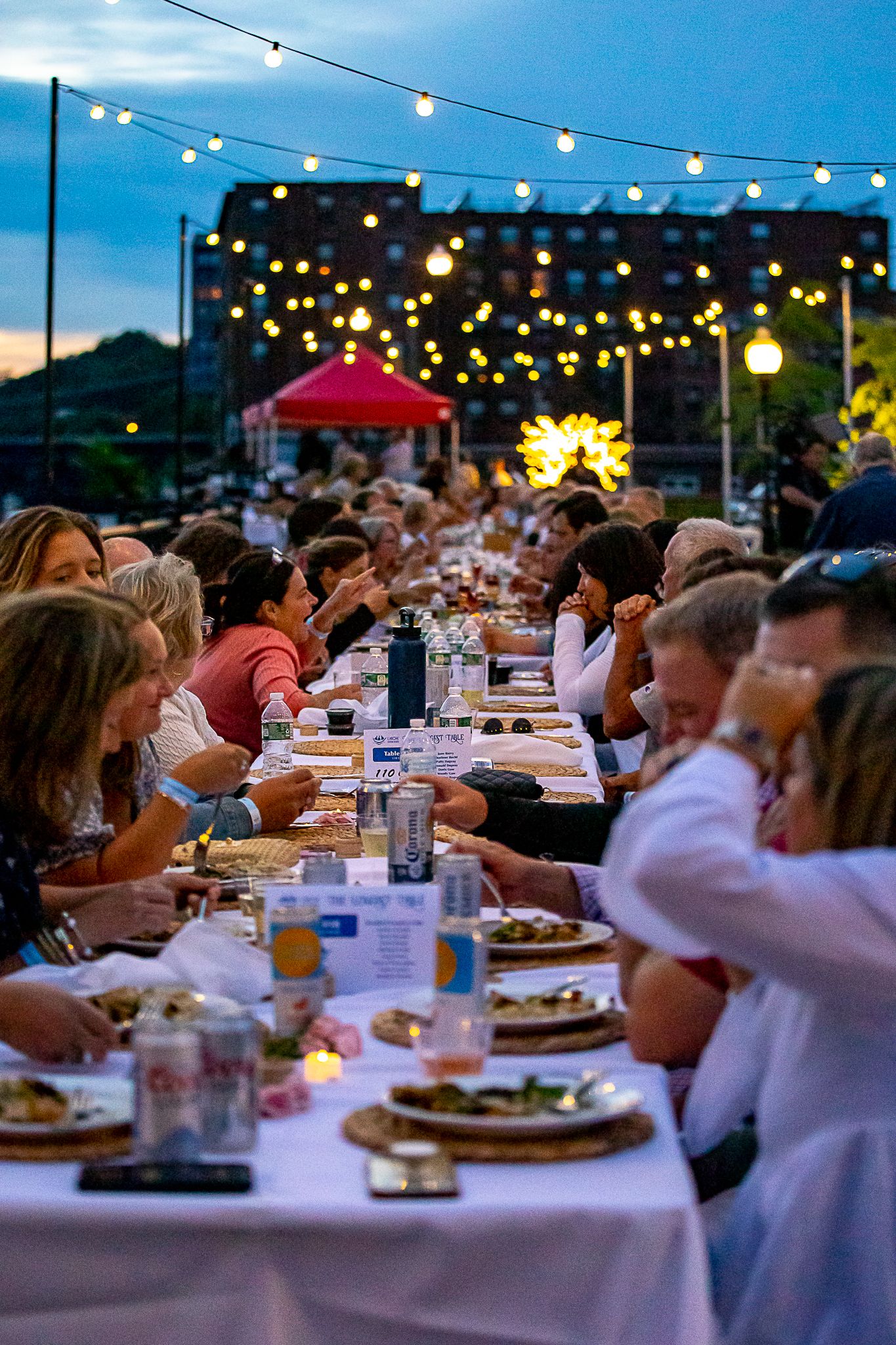 Outdoor evening dinner party with string lights, long tables of guests dining al fresco at dusk in an urban setting.
