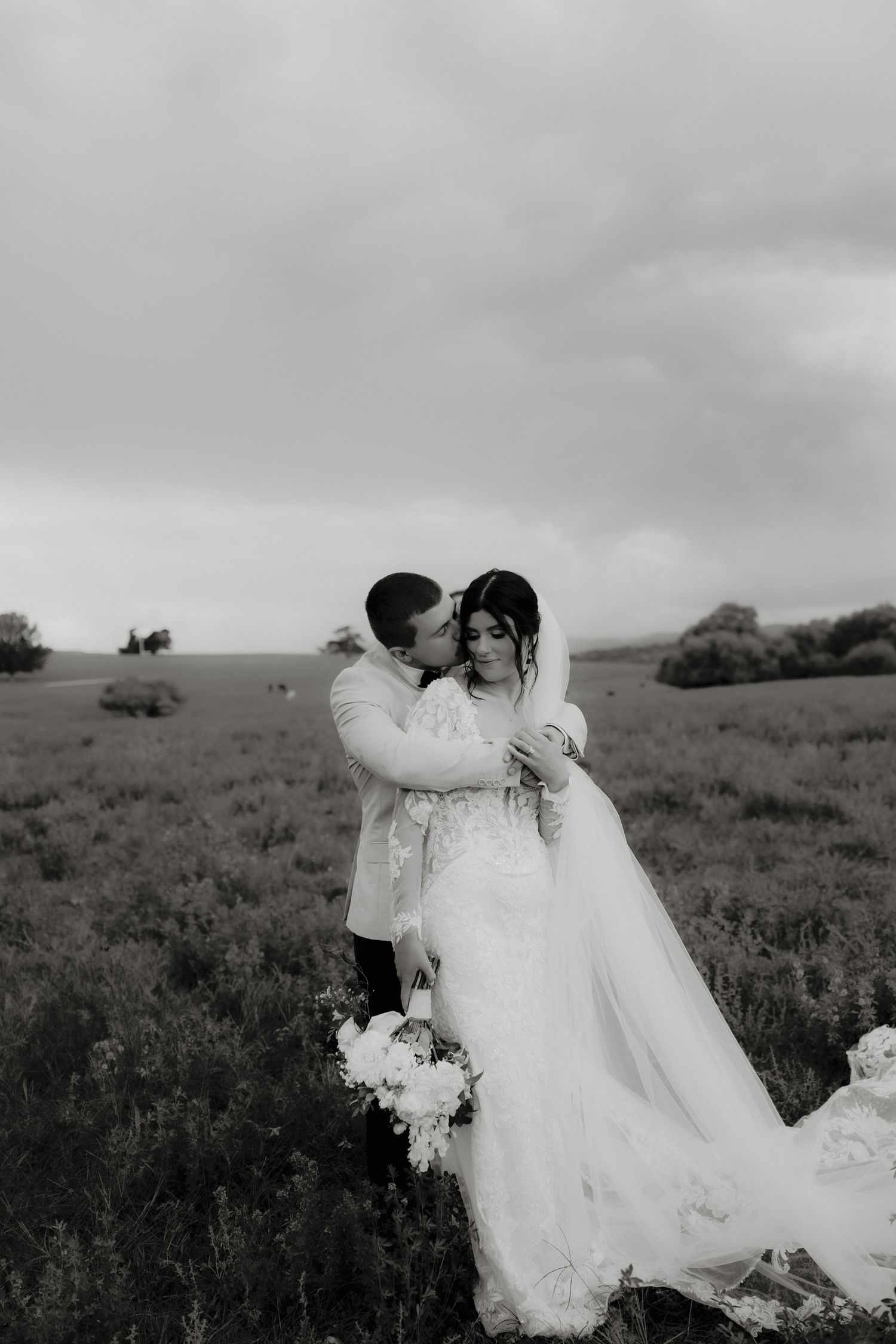 Black and white photo of a groom kissing bride on forehead in a field, bride holding white bouquet in wedding dress.