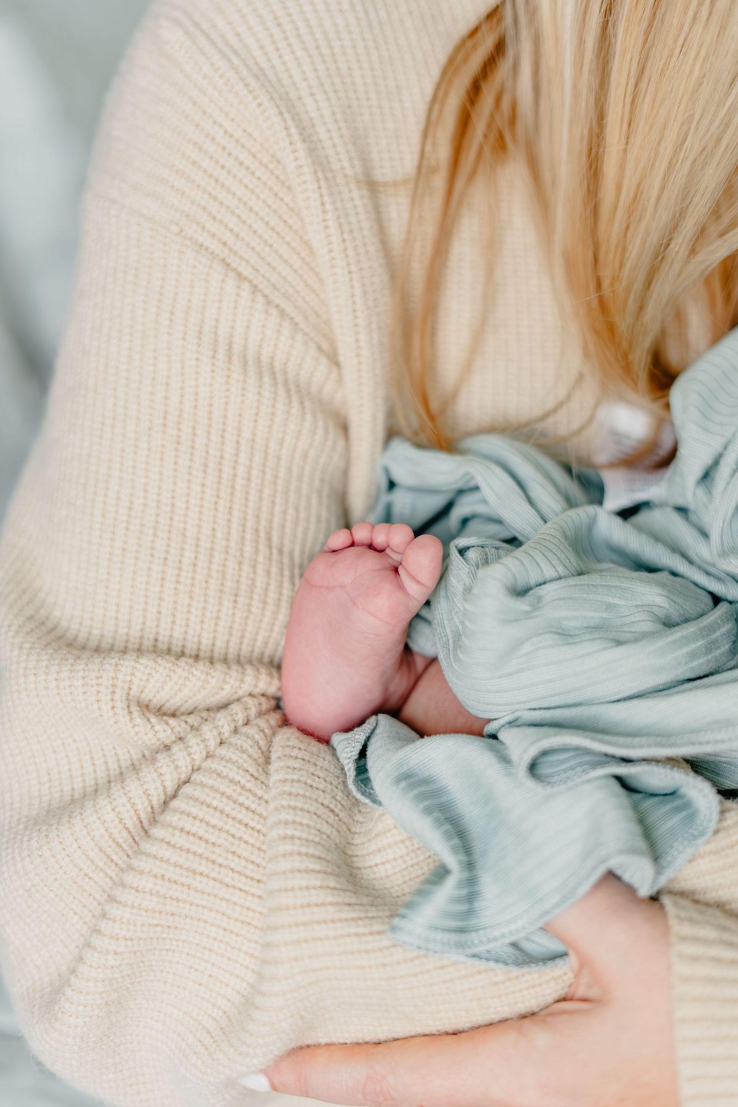 A mother in a beige knit sweater cradles her newborn baby wrapped in a soft mint green blanket.