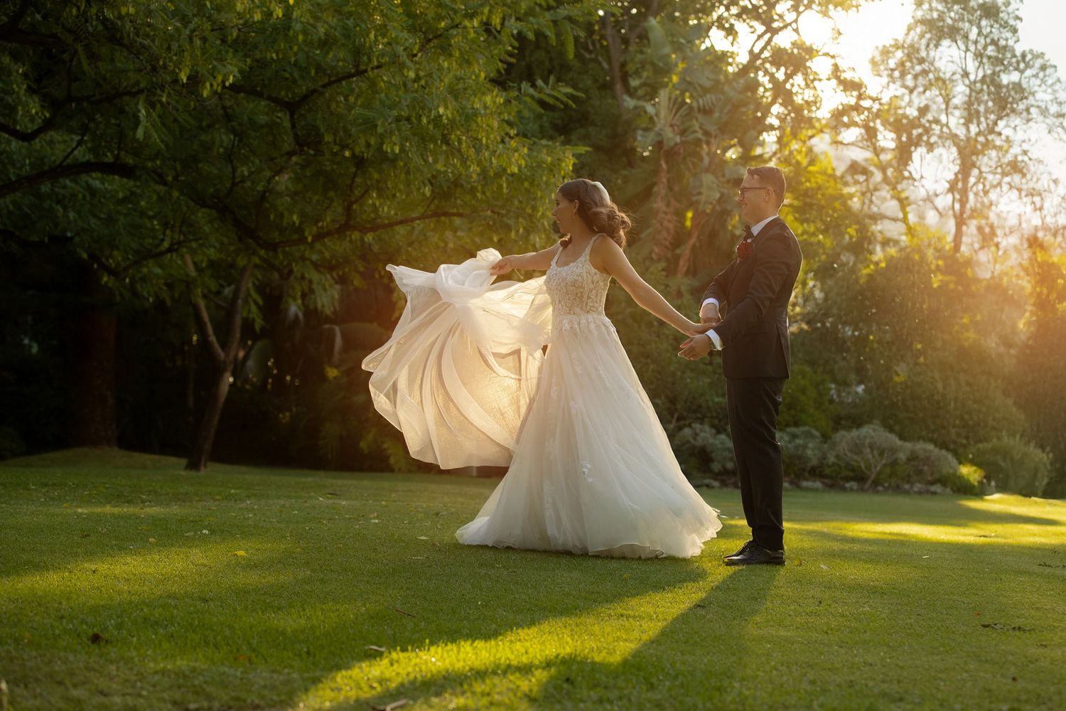 Bride in flowing white gown twirling with groom in black tuxedo during golden hour outdoor wedding ceremony.