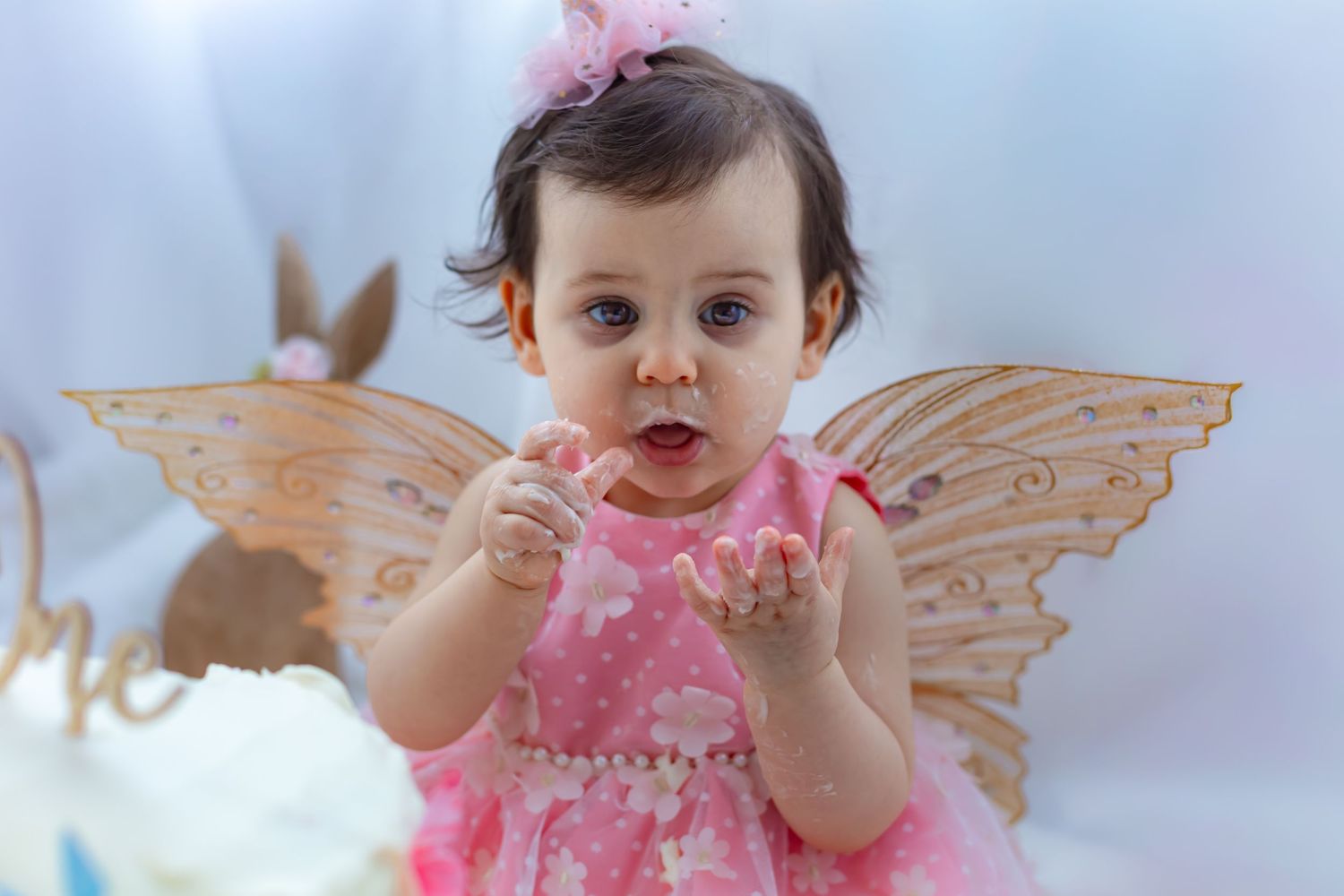 Baby girl in pink floral dress with fairy wings and bow headband celebrates first birthday with cake smash photo.