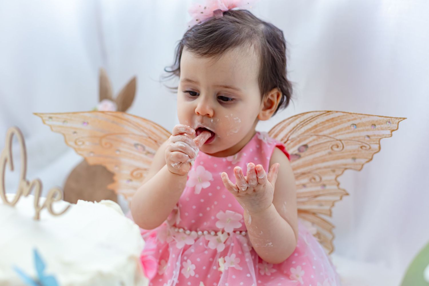 Baby girl in pink dress with fairy wings eating cake at her first birthday party celebration.