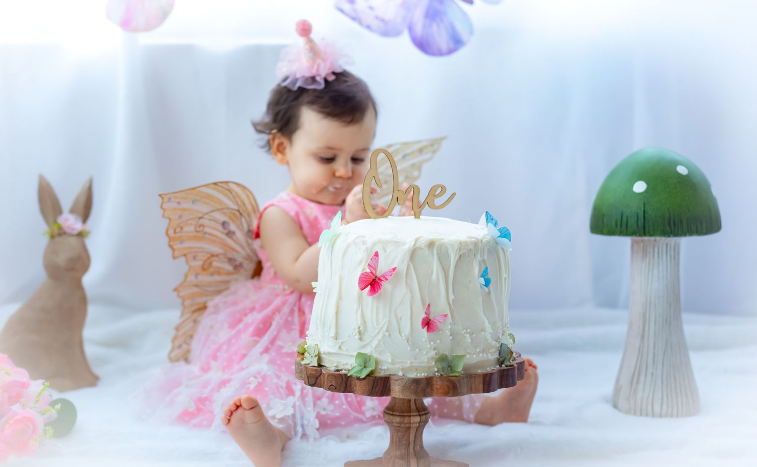 Baby girl in pink butterfly dress touches her first birthday cake beside decorative mushroom and bunny props.