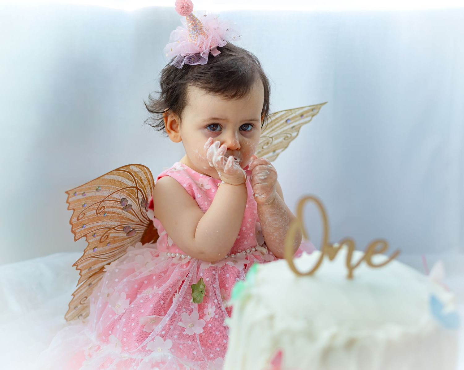 Baby girl in pink dress with fairy wings celebrates her first birthday smashing cake, wearing a pink crown.