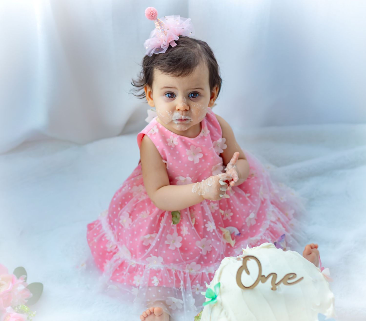 Baby girl in pink floral dress celebrates first birthday with cake smash, wearing a pink crown headband.