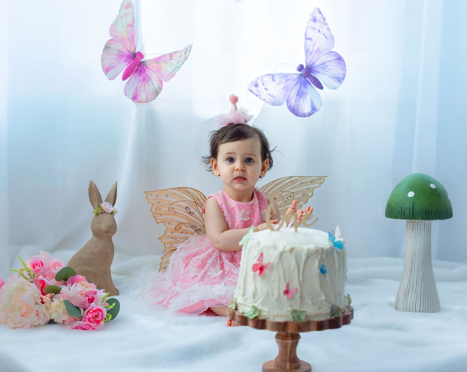 Baby girl in pink fairy dress with wings sits beside a white cake, butterflies, bunny, and flowers in a whimsical setup.