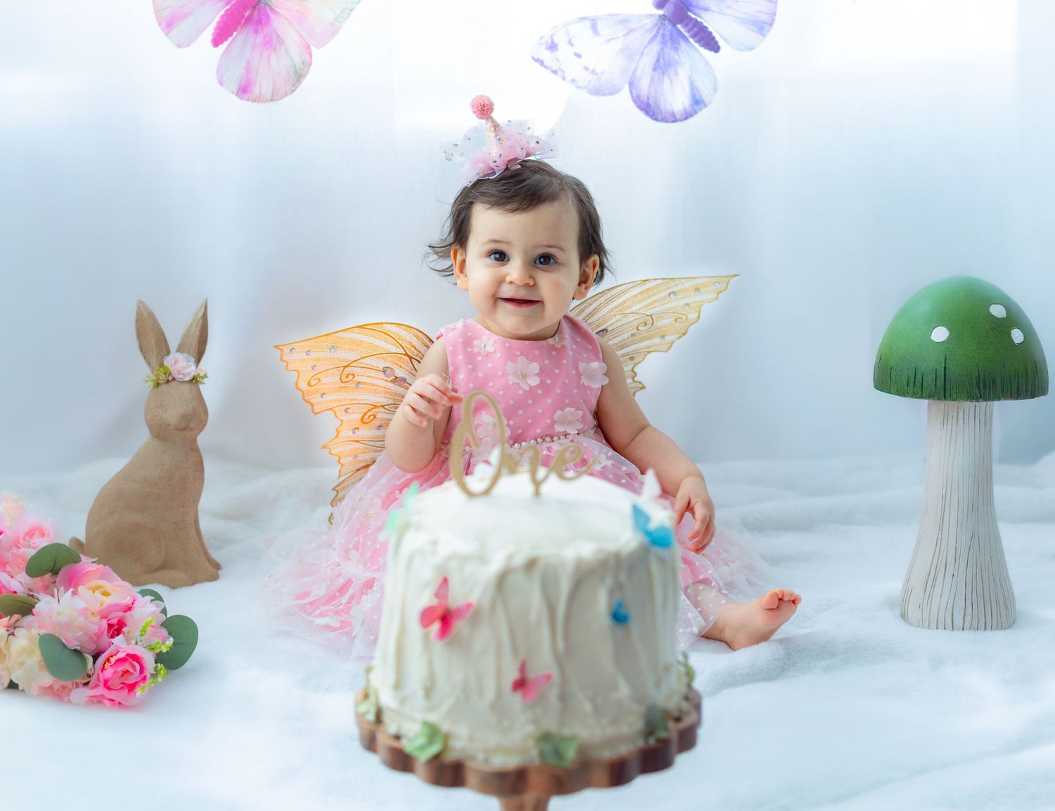 Baby girl in pink fairy dress with butterfly wings sits before a white cake, surrounded by mushroom and bunny decor.