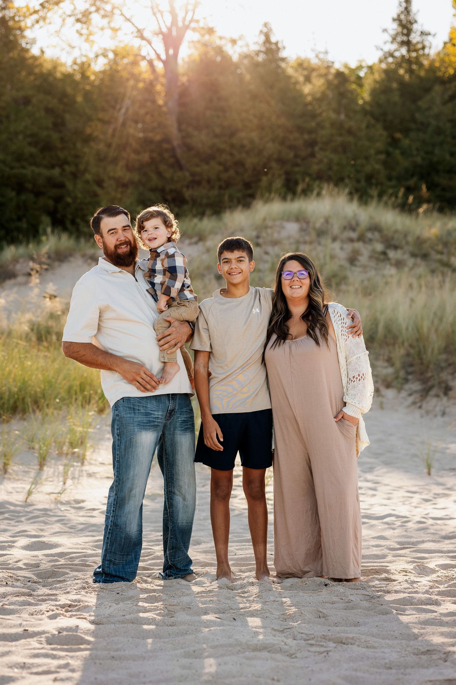 Family of four posing on sandy beach dunes at golden hour, father holding toddler, pregnant mother in maxi dress.