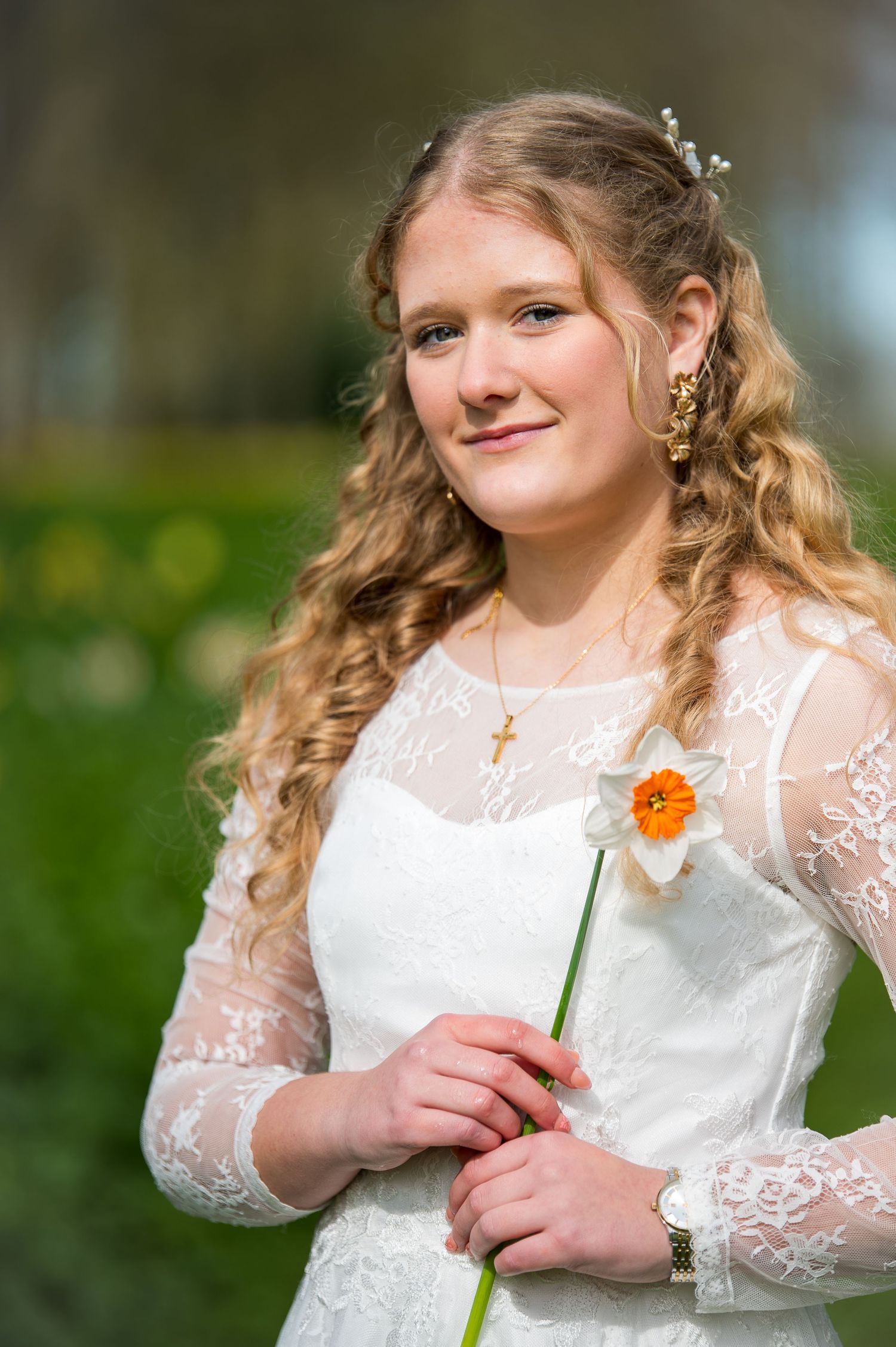 Young woman in white lace dress holding a daffodil, wearing a cross necklace, with curly blonde hair outdoors.