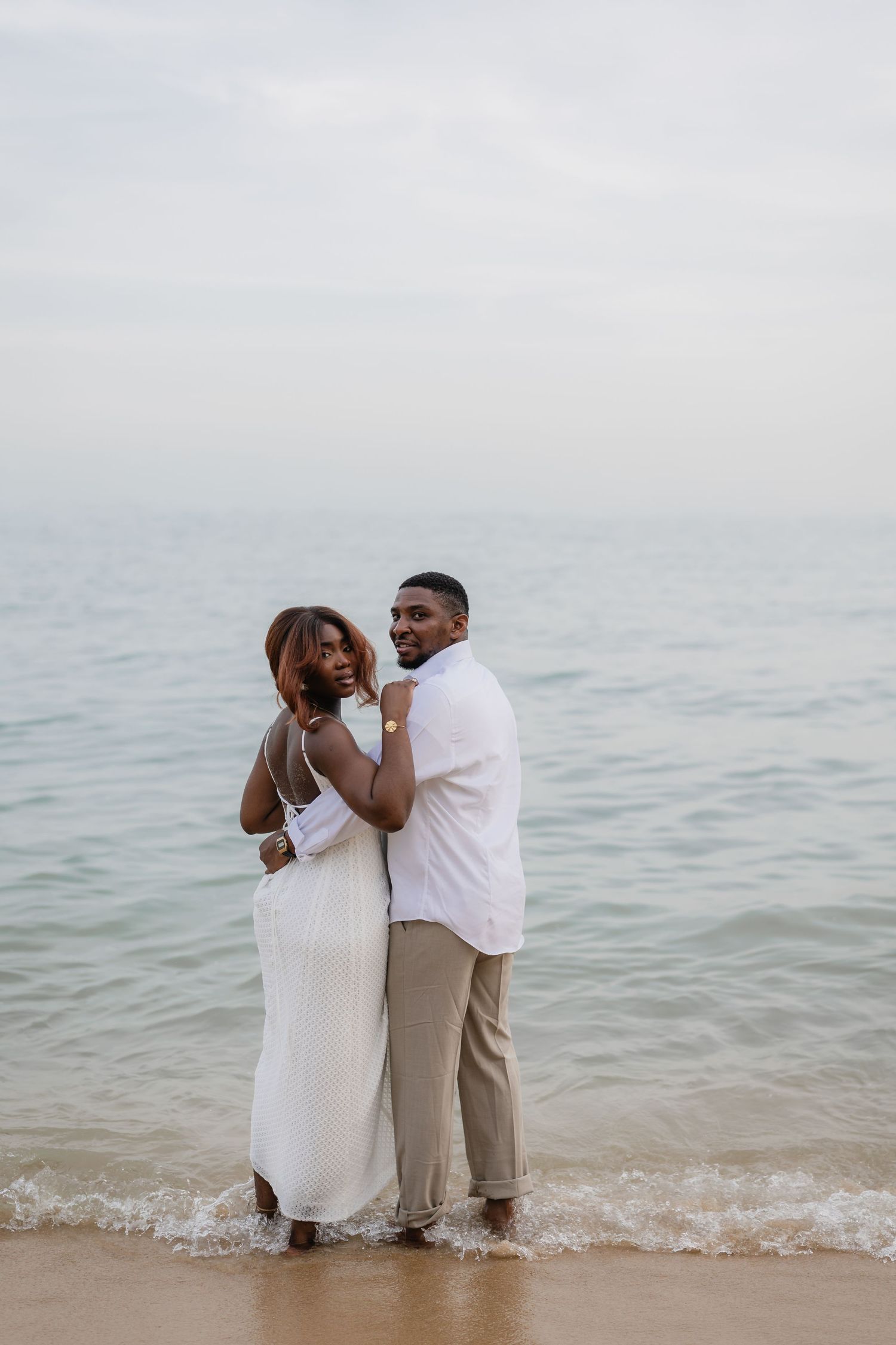 A couple in white outfits embracing on a sandy beach with calm ocean waves at their feet.