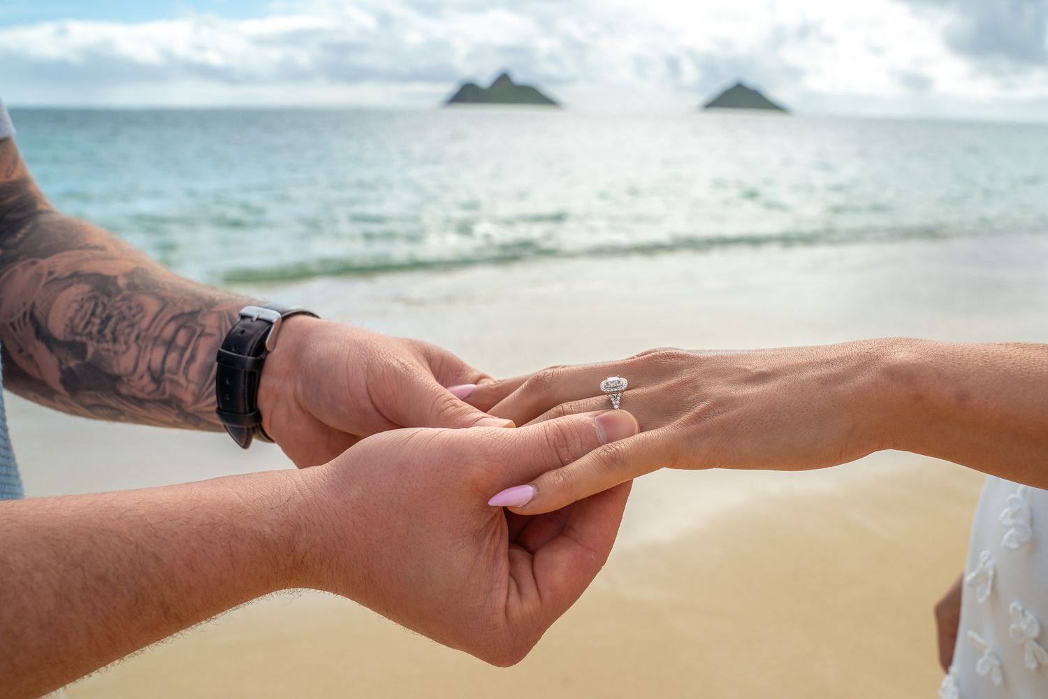 Groom with tattoo placing engagement ring on bride's finger on a sandy beach with island views.