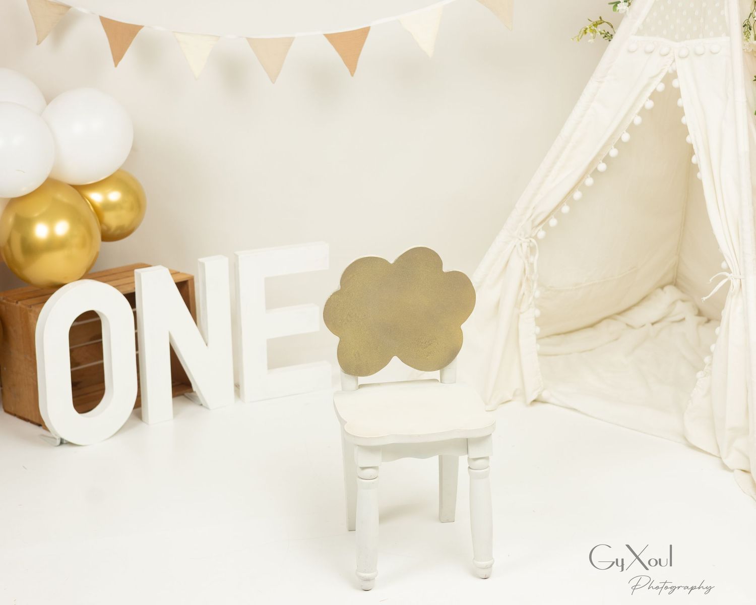 First birthday photo setup with ONE letters, white teepee, gold balloons, and a small chair with cloud cutout.